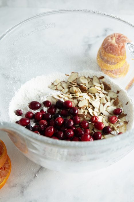 A clear glass bowl containing flour, sliced almonds, and red cranberries, with an orange juicer and pressed orange halves in the background on a marble surface.