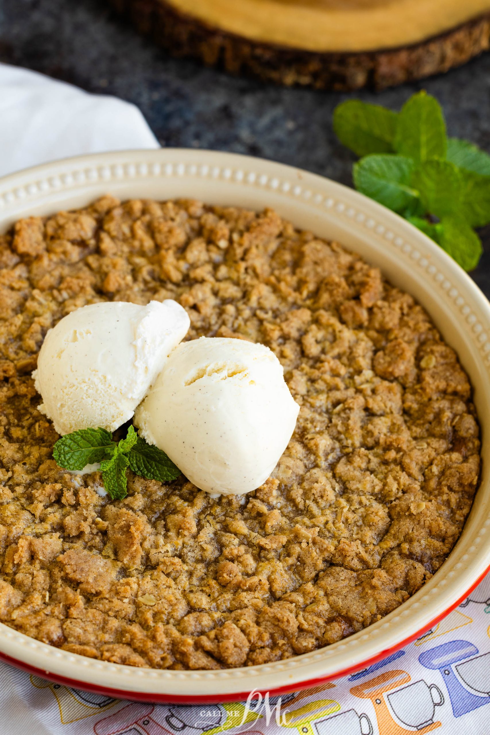 A round dish with a crumbly dessert topped with two scoops of vanilla ice cream, garnished with mint leaves.