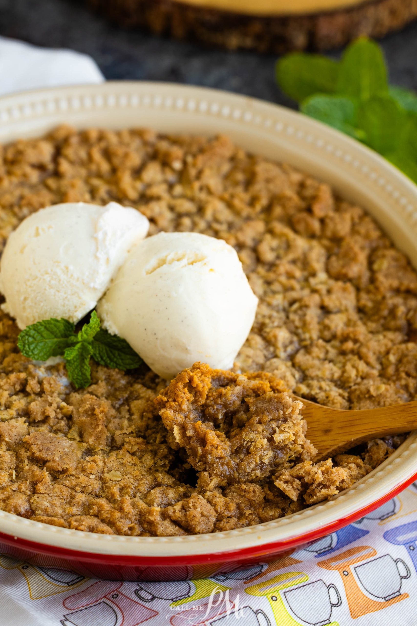 A round baking dish filled with apple crisp topped with two scoops of vanilla ice cream and a sprig of mint. A wooden spoon is scooping a portion.