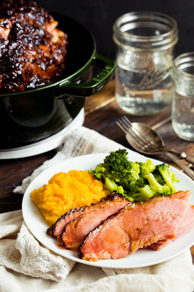 Plate of sliced glazed ham with mashed squash and steamed broccoli, next to a pot with more ham and two mason jars on a wooden table.