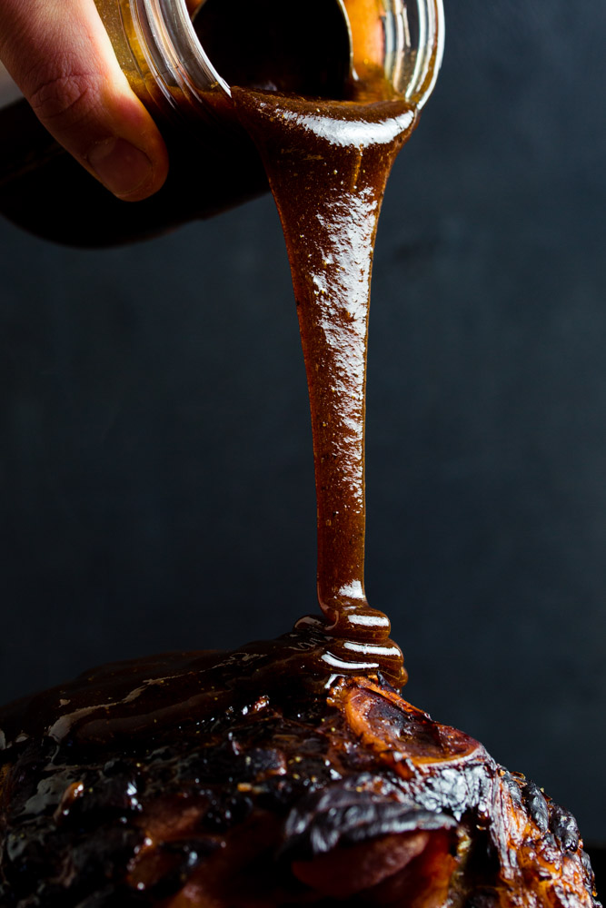 Barbecue sauce being poured from a jar onto a grilled meat dish against a dark background.