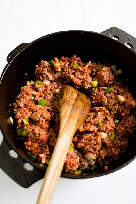 Wooden spoon stirring seasoned ground meat with diced vegetables in a black pot.