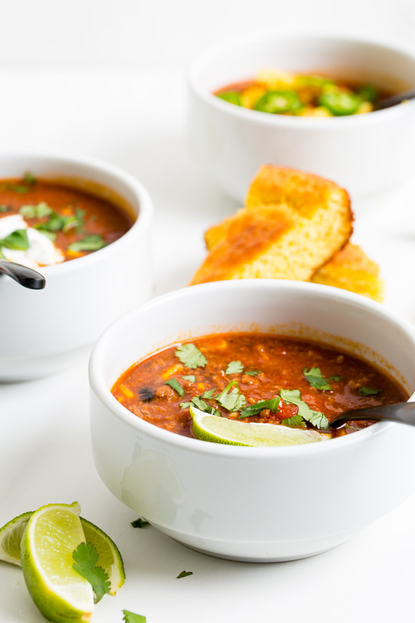 Three white bowls of Dutch Oven Taco Chili garnished with cilantro and lime wedges, served with cornbread slices on a white background.