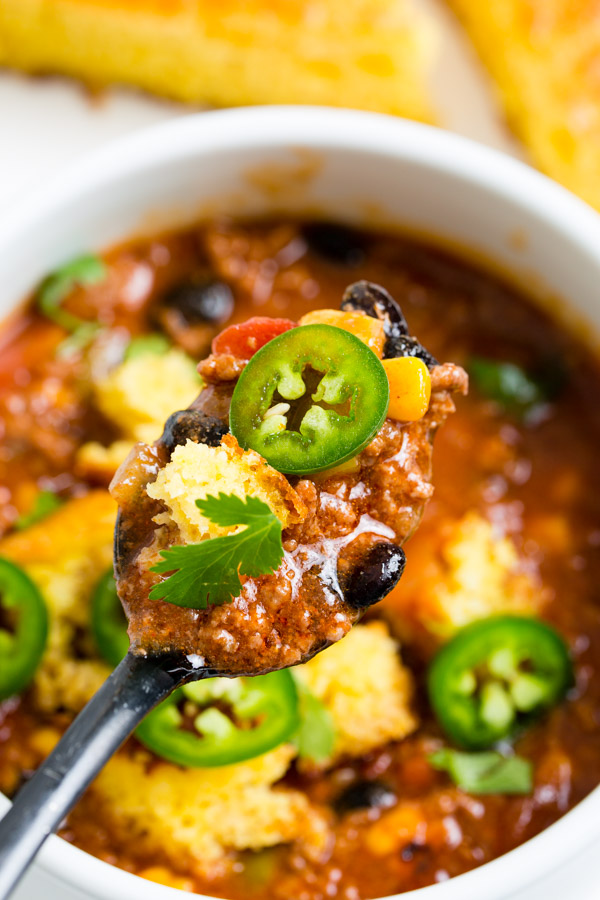 A spoonful of chili with black beans, corn, jalapeño slices, and cilantro is held above a bowl containing the same ingredients, with cornbread pieces visible.