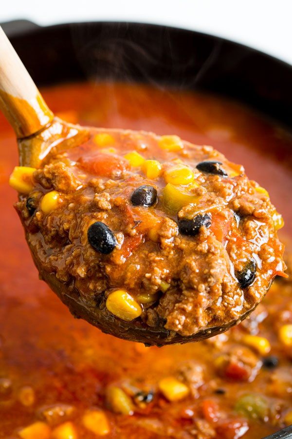 A ladle of hearty Dutch Oven Taco Chili with corn, black beans, and ground meat is held above a pot of red chili. Steam rises from the hot dish.