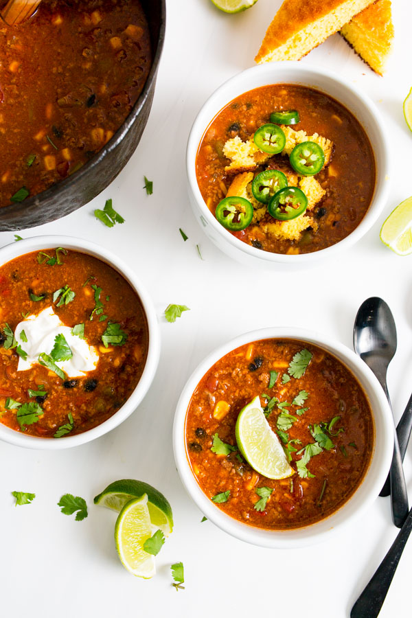 Three bowls of Dutch Oven Taco Chili with various toppings like lime, sour cream, cilantro, jalapeños, and cornbread on a white surface. A pot of chili and lime wedges are nearby.