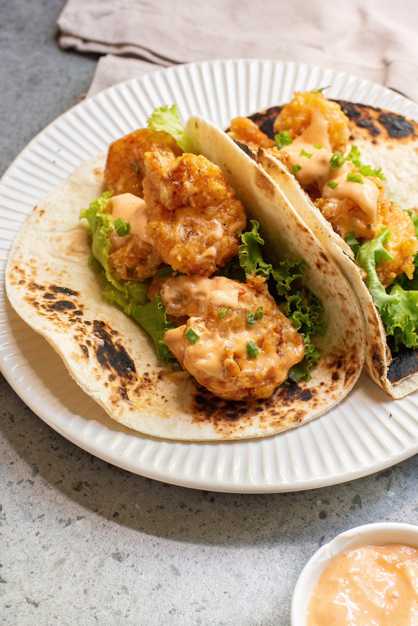 Two crispy shrimp tacos with lettuce and sauce on a white plate, accompanied by a small bowl of sauce.