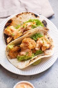 Two tacos filled with crispy fried shrimp, lettuce, and a drizzle of sauce, served on a white plate.