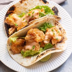 Two tacos filled with crispy fried shrimp, lettuce, and a drizzle of sauce, served on a white plate.