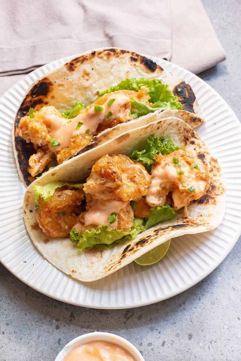 Two tacos filled with crispy fried shrimp, lettuce, and a drizzle of sauce, served on a white plate.