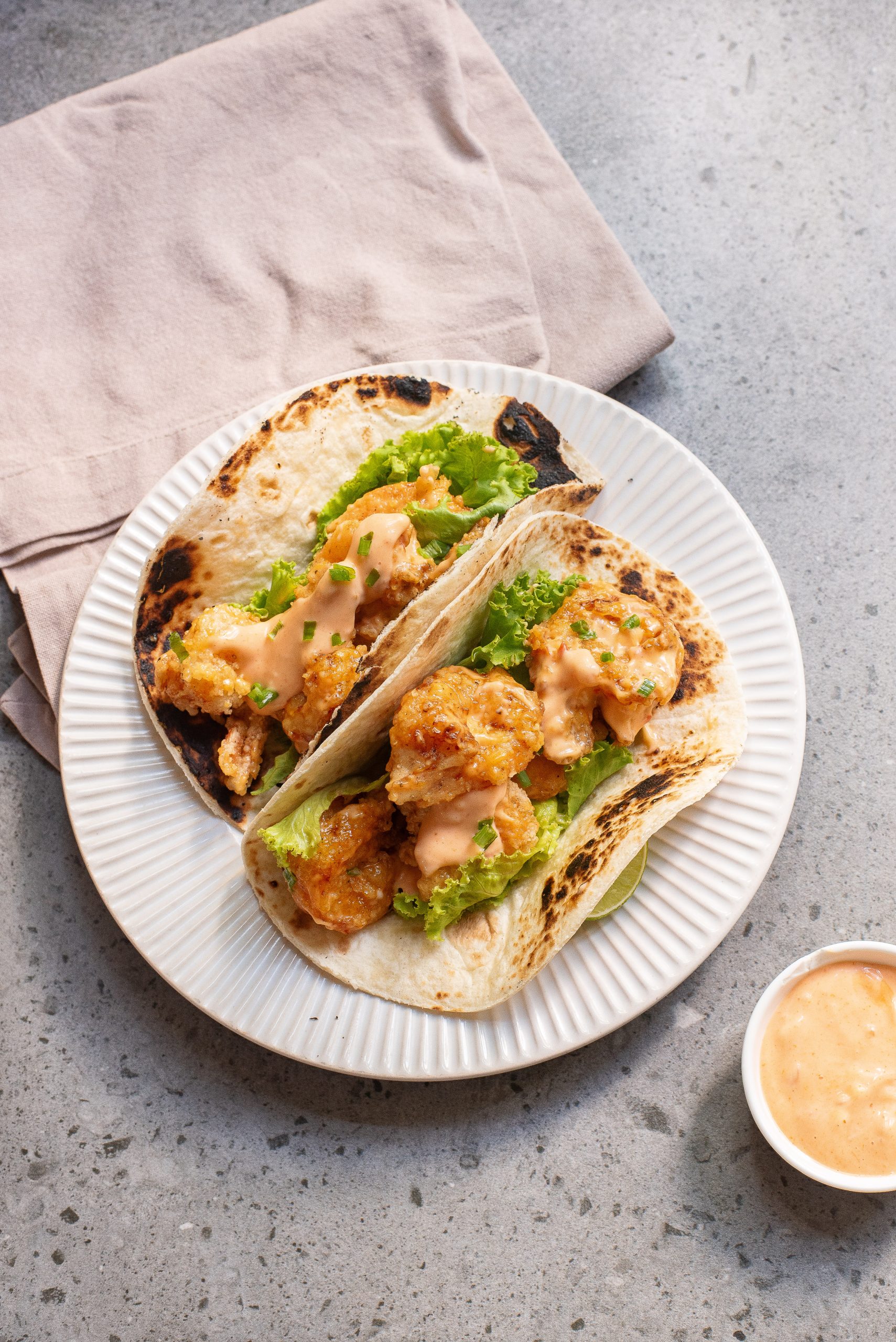 Two grilled tortillas filled with fried chicken pieces, lettuce, and creamy sauce on a white plate with a dip bowl and napkin on the side.