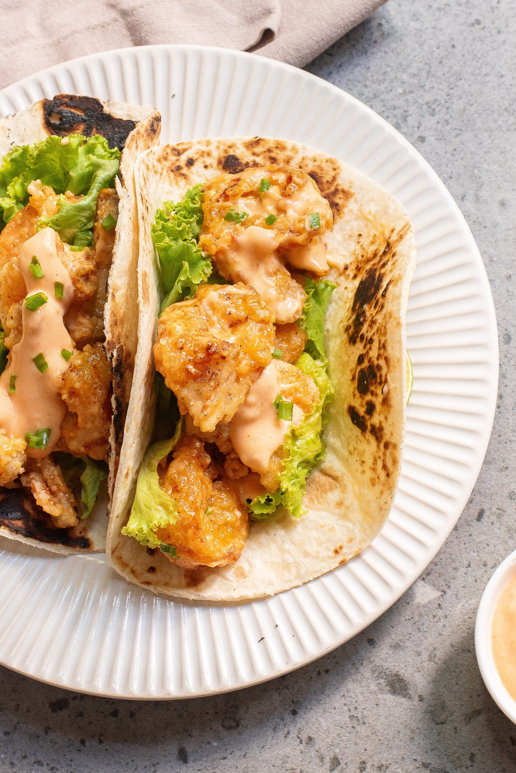 Two soft-shell tacos on a white plate, each filled with fried shrimp, lettuce, and drizzled with a creamy sauce.