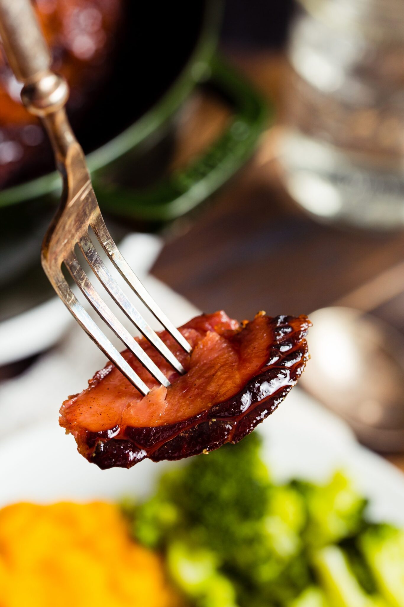 A fork holds up a slice of glazed ham. Broccoli and mashed sweet potatoes are visible in the background.