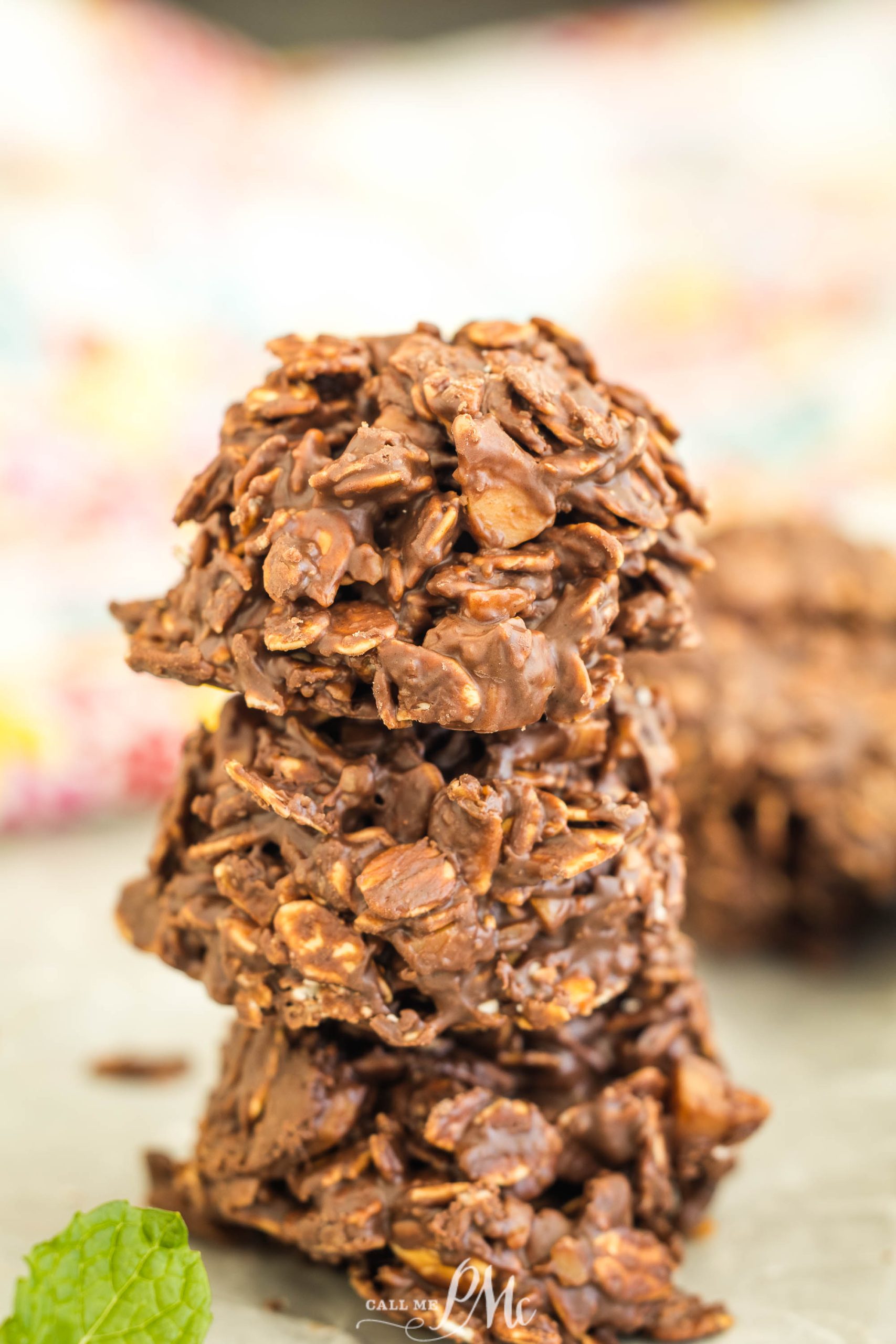 A stack of three chocolate oatmeal clusters on a surface, with a blurred background and a mint leaf nearby.