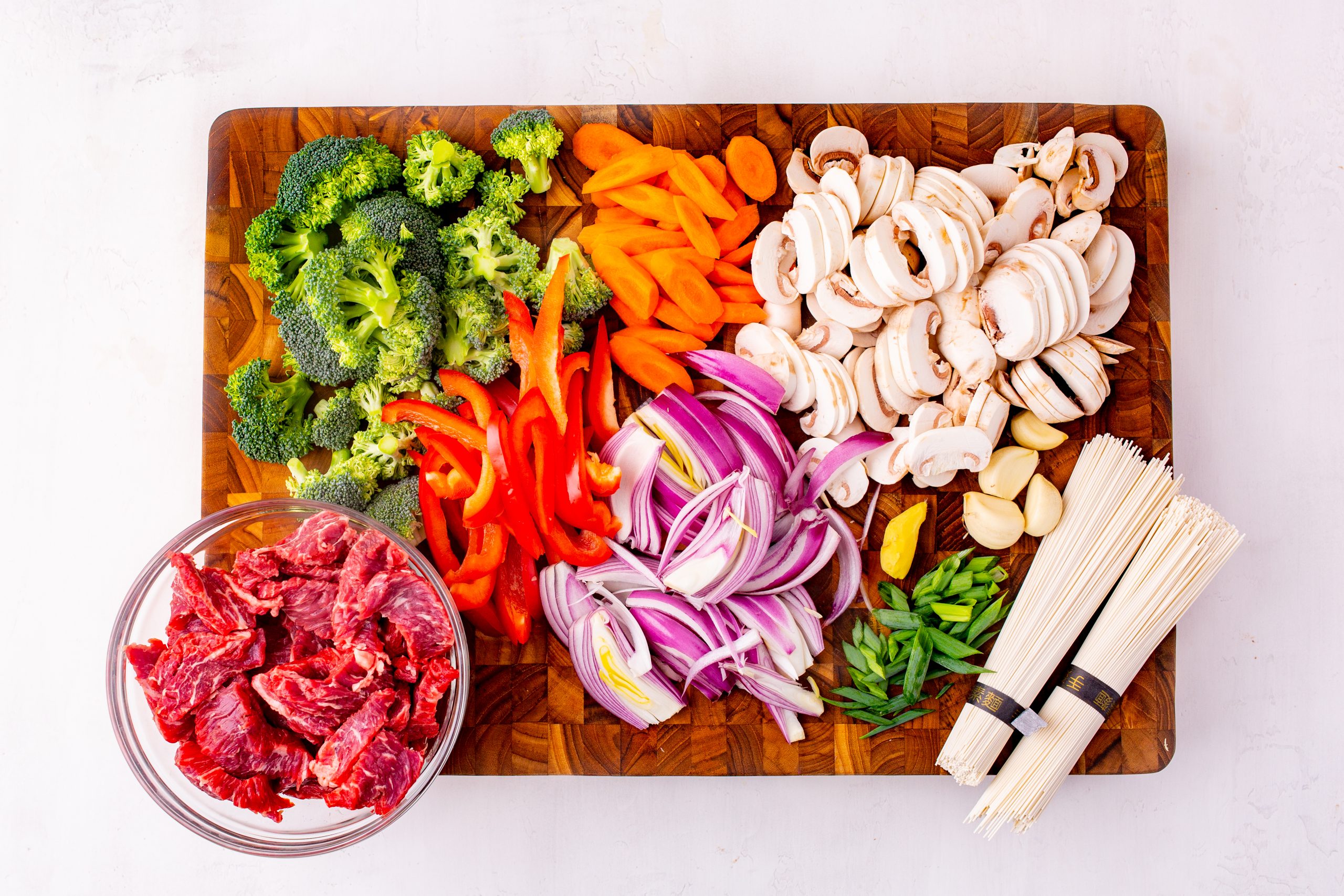 Ingredients arranged on a cutting board: sliced beef in a bowl, broccoli, carrots, red peppers, red onions, mushrooms, garlic cloves, green onions, and bundles of noodles.