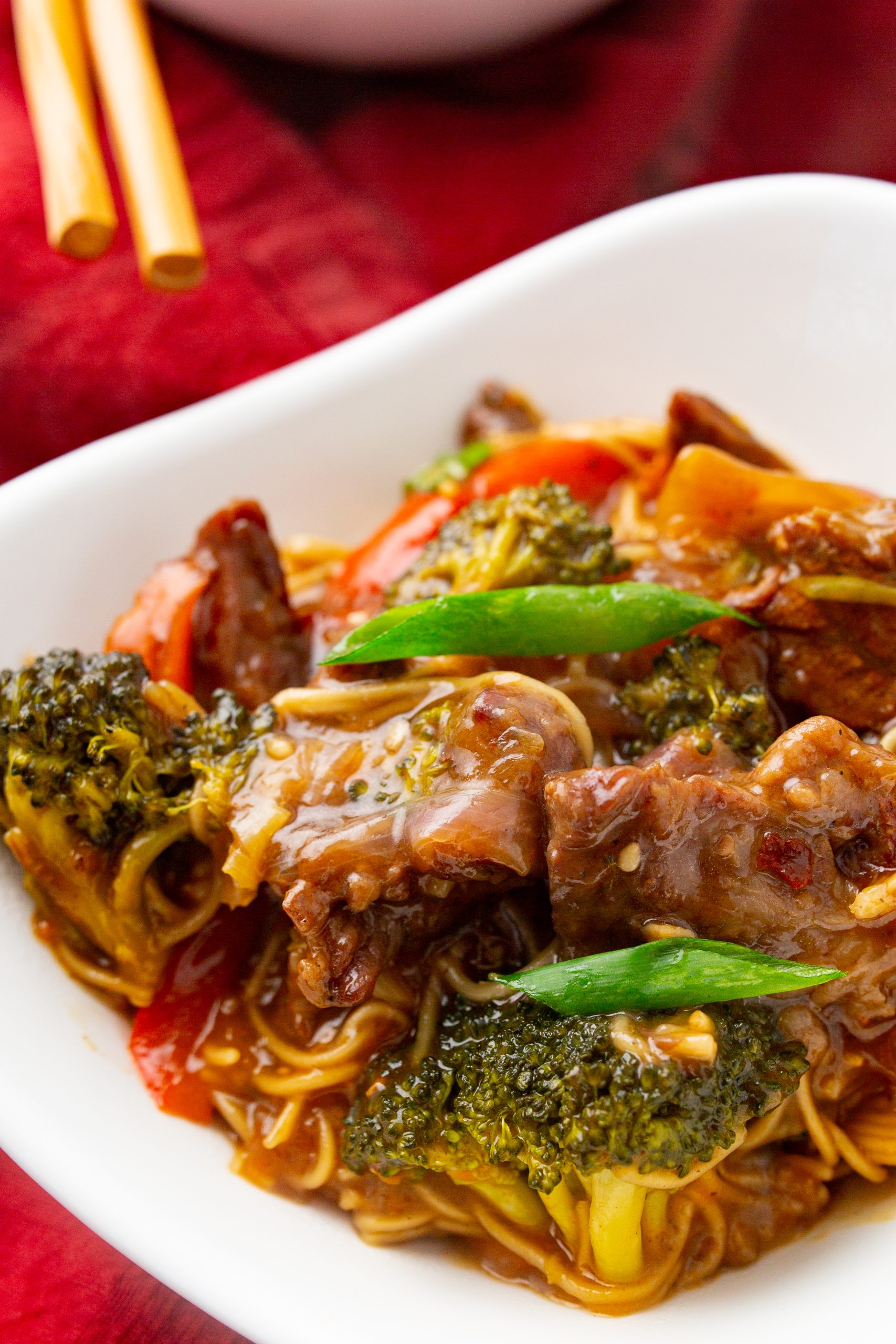 A bowl of beef stir-fry with broccoli, bell peppers, and green onions, served over noodles in a white dish. Chopsticks rest on the table beside it.