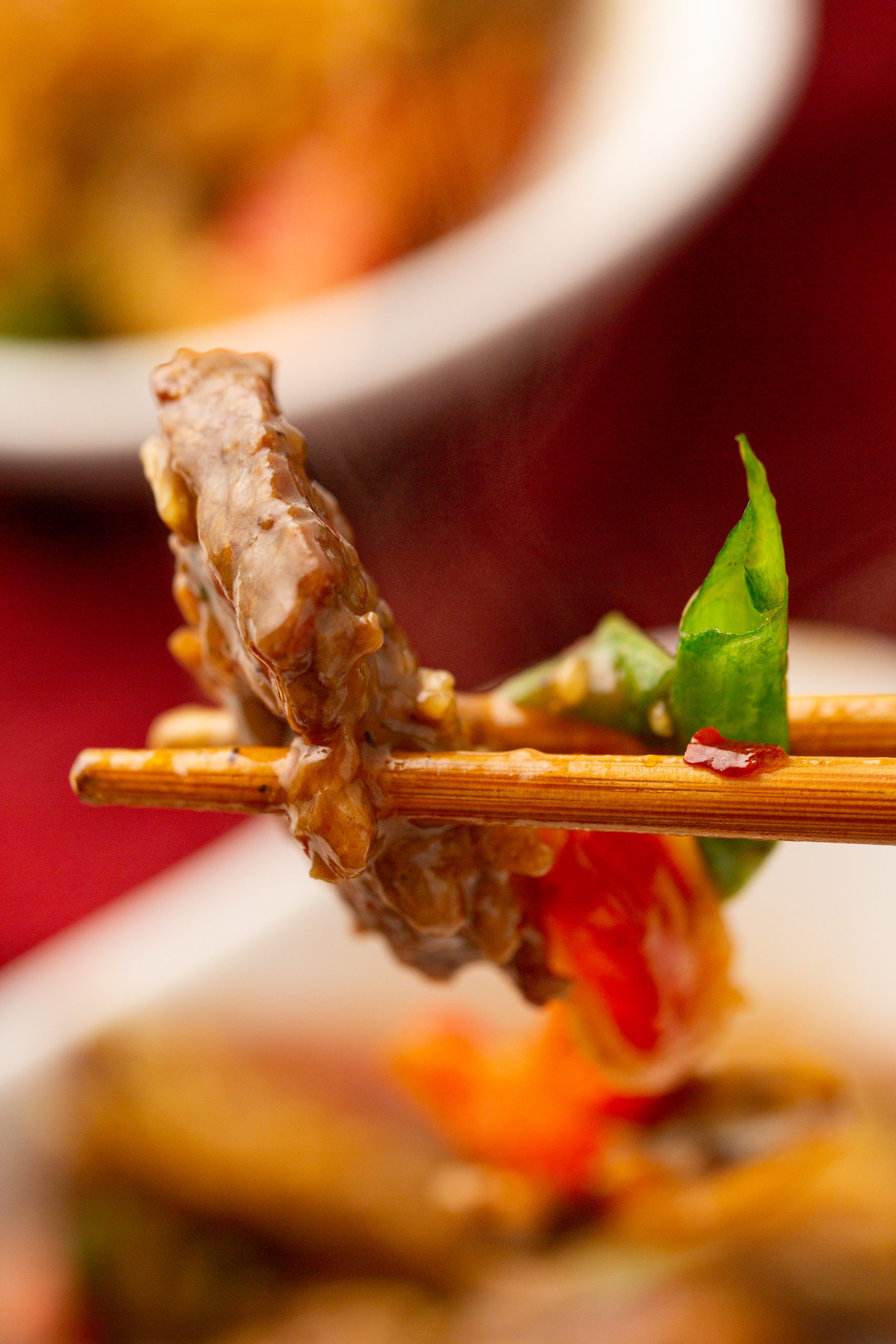 Close-up of chopsticks holding a piece of stir-fried beef with vegetables over a blurred background.