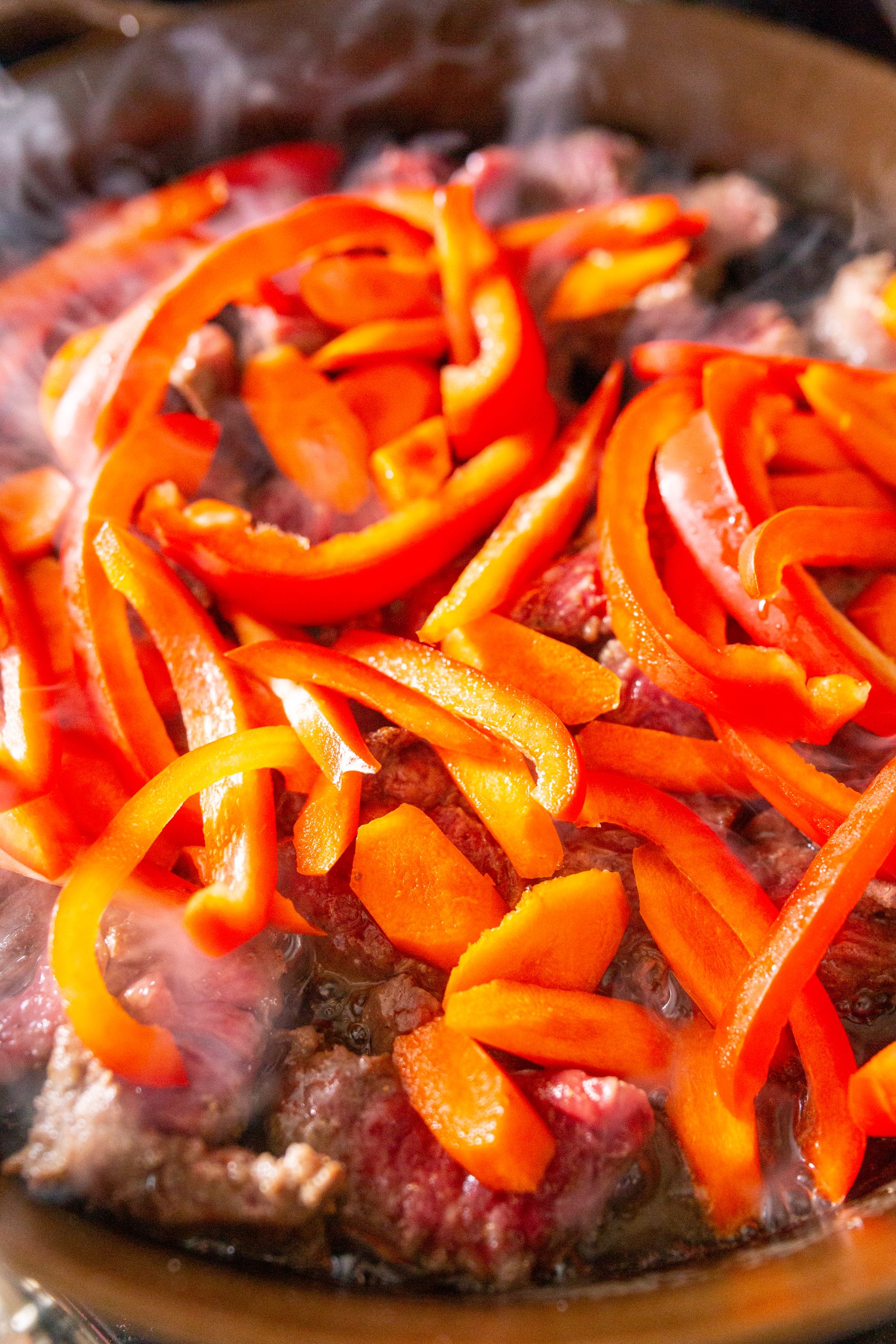 Sliced red bell peppers and pieces of beef cooking in a pan with visible steam.