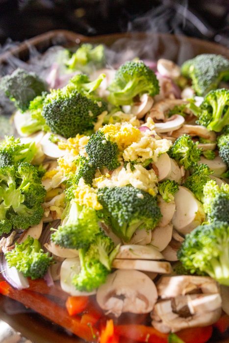 Close-up of chopped broccoli, mushrooms, red onions, and garlic in a pan, surrounded by steam.