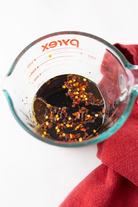 A glass measuring cup with dark liquid and red pepper flakes on top, placed on a red cloth.