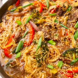 A pan filled with stir-fried noodles, beef slices, red peppers, broccoli, mushrooms, green onions, and sesame seeds in a savory sauce.