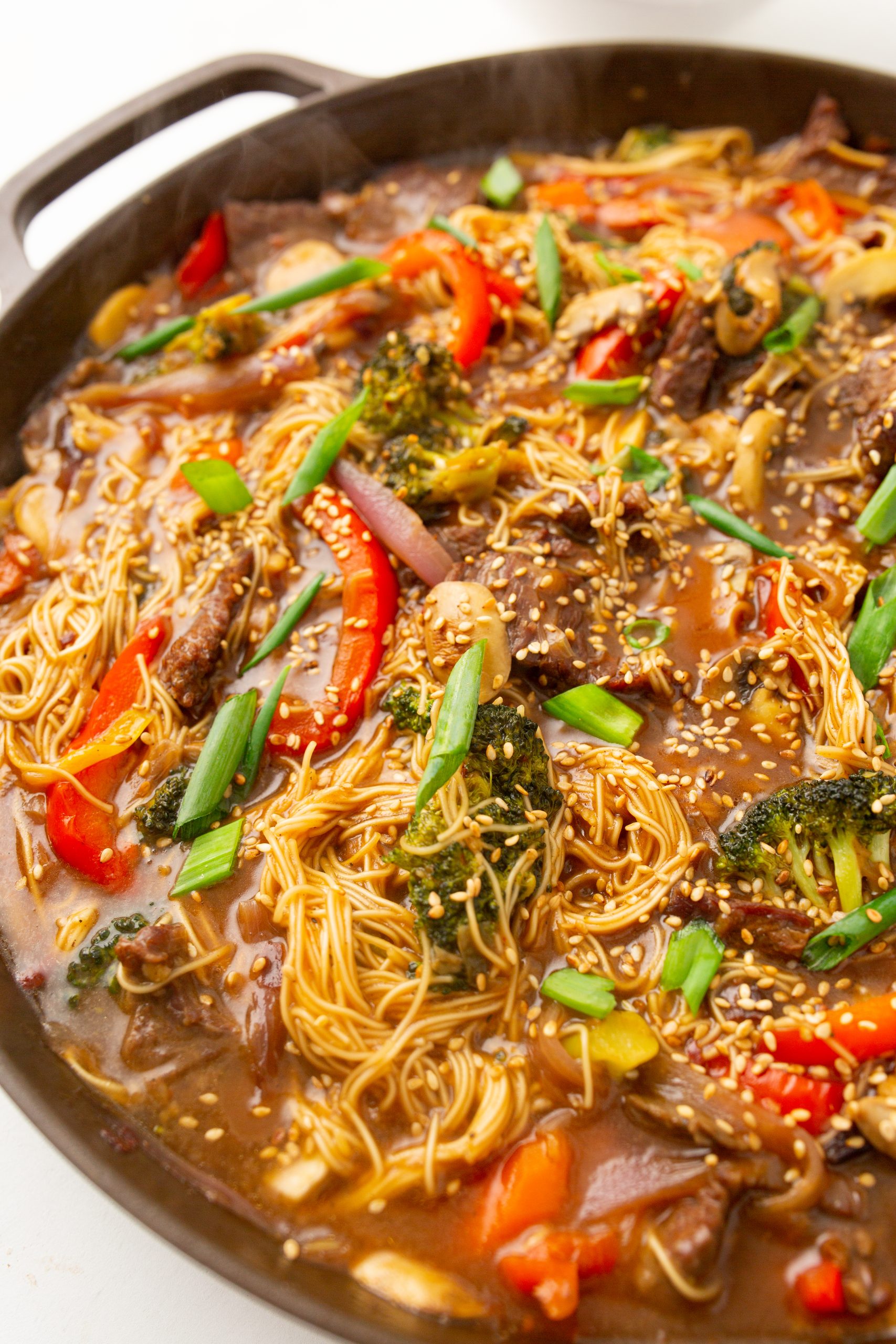 A pan filled with stir-fried noodles, beef slices, red peppers, broccoli, mushrooms, green onions, and sesame seeds in a savory sauce.