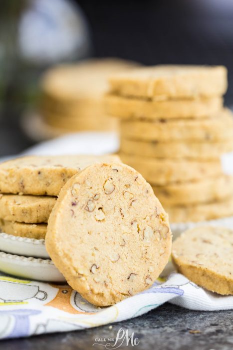 A stack of round pecan shortbread cookies on a white plate, with one cookie leaning in front, placed on a patterned cloth.