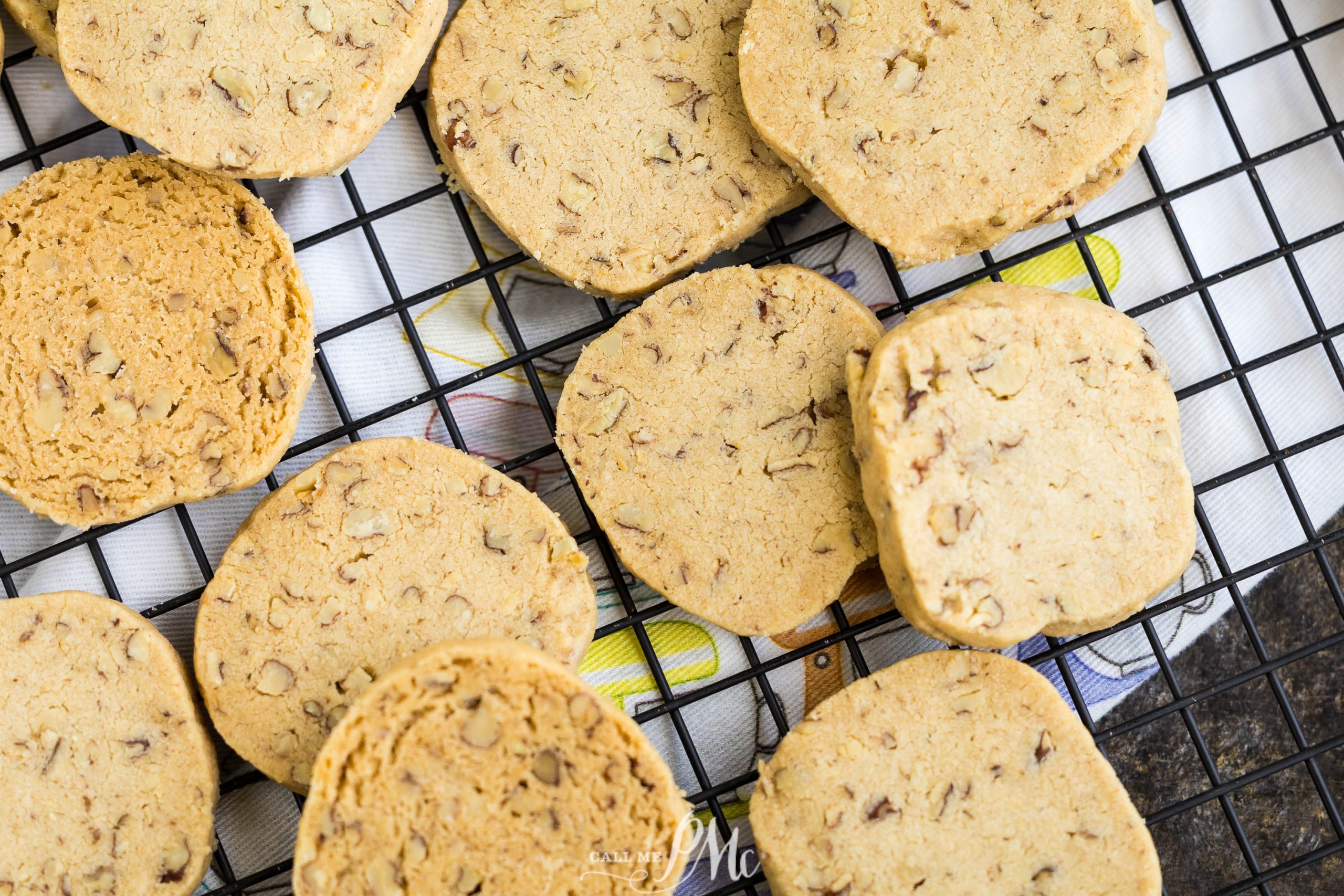 A variety of round and square-shaped cookies with nuts rest on a cooling rack.