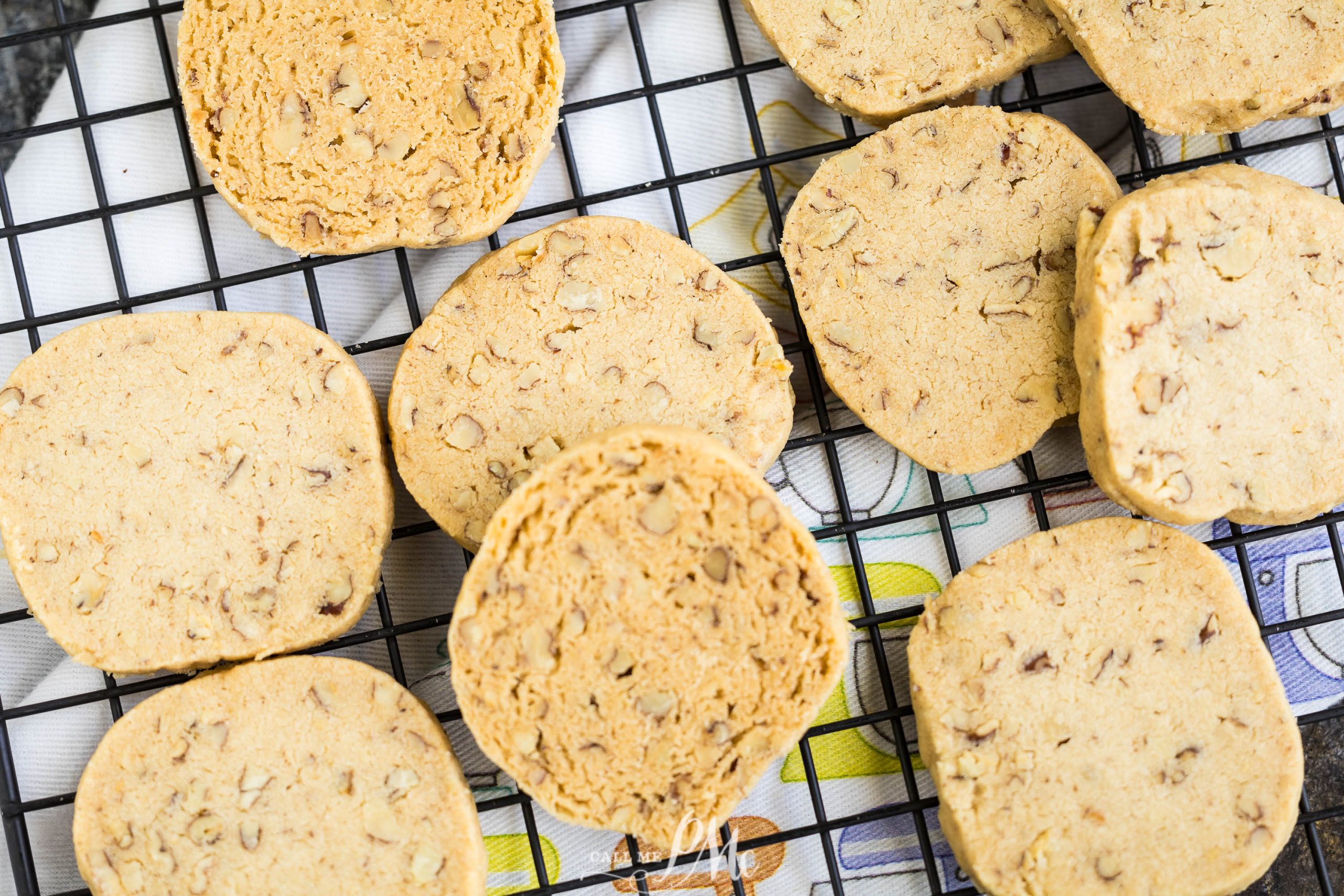 Several Pecan Sandies Recipe with nut pieces rest on a black wire cooling rack over a patterned cloth.