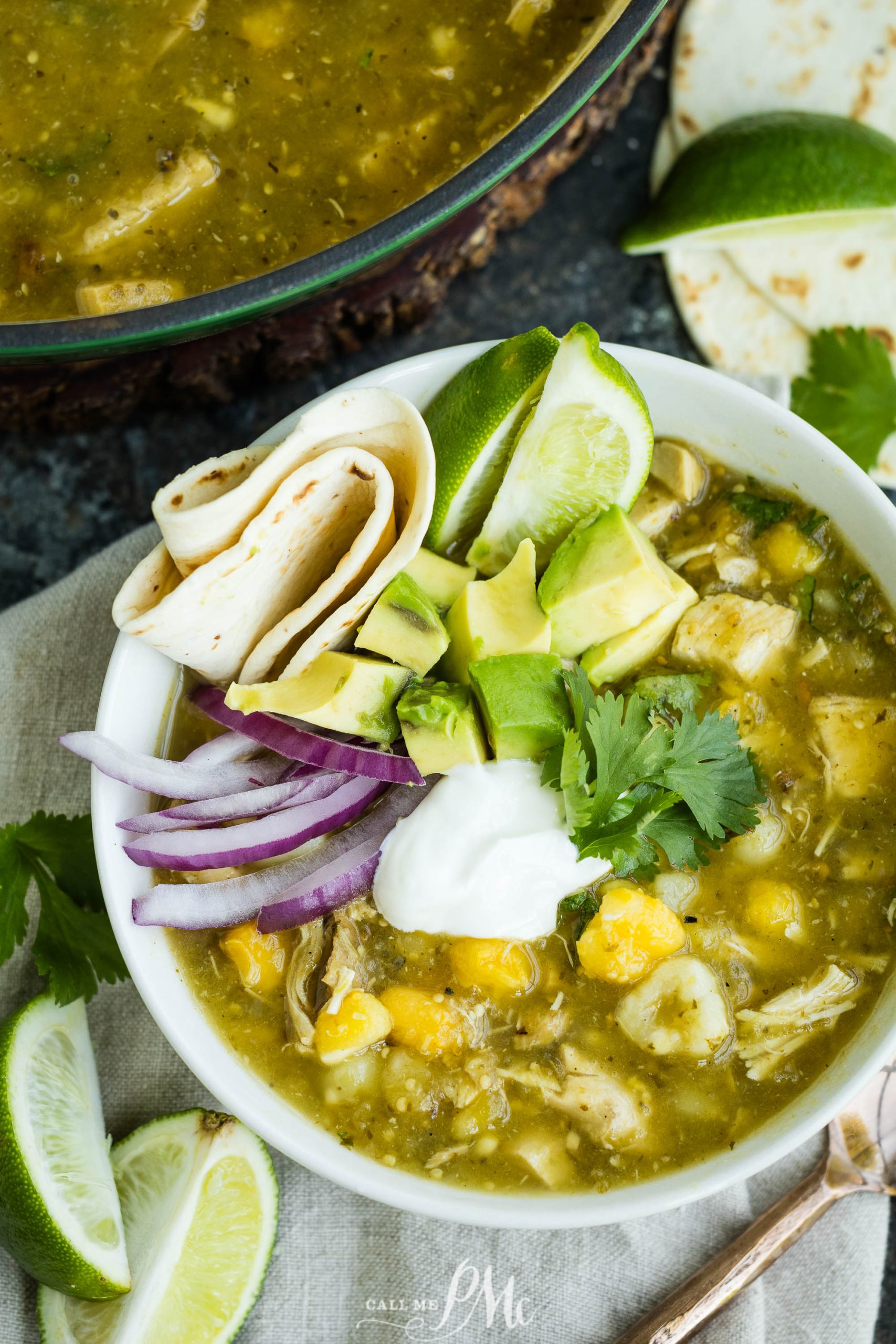 A bowl of chicken chili verde soup garnished with sliced avocado, red onion, sour cream, cilantro, and lime wedges, with a tortilla on the side.