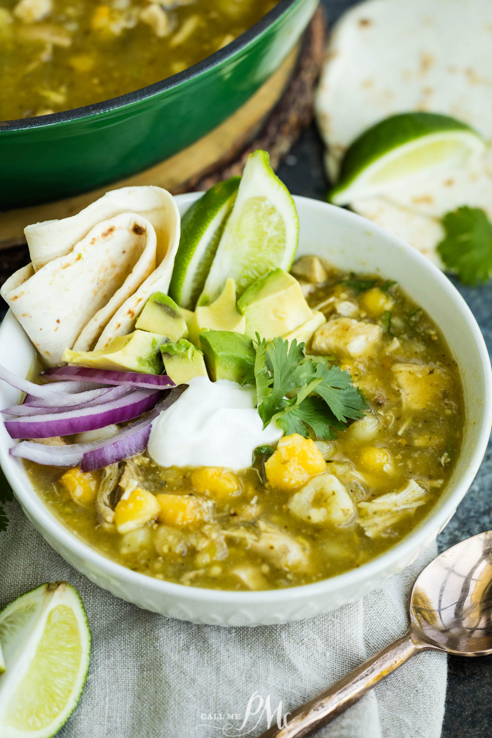 A bowl of green chili stew with chicken, potatoes, avocado slices, lime wedges, sour cream, onion, and flour tortillas beside it.