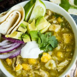 Bowl of chicken chili verde topped with avocado, sour cream, red onion, cilantro, and a lime wedge. A tortilla is rolled on the side.