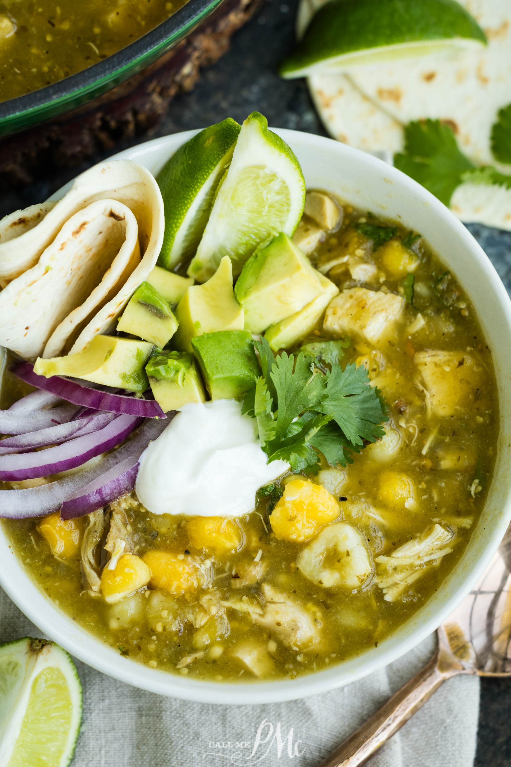 Bowl of chicken chili verde topped with avocado, sour cream, red onion, cilantro, and a lime wedge. A tortilla is rolled on the side.