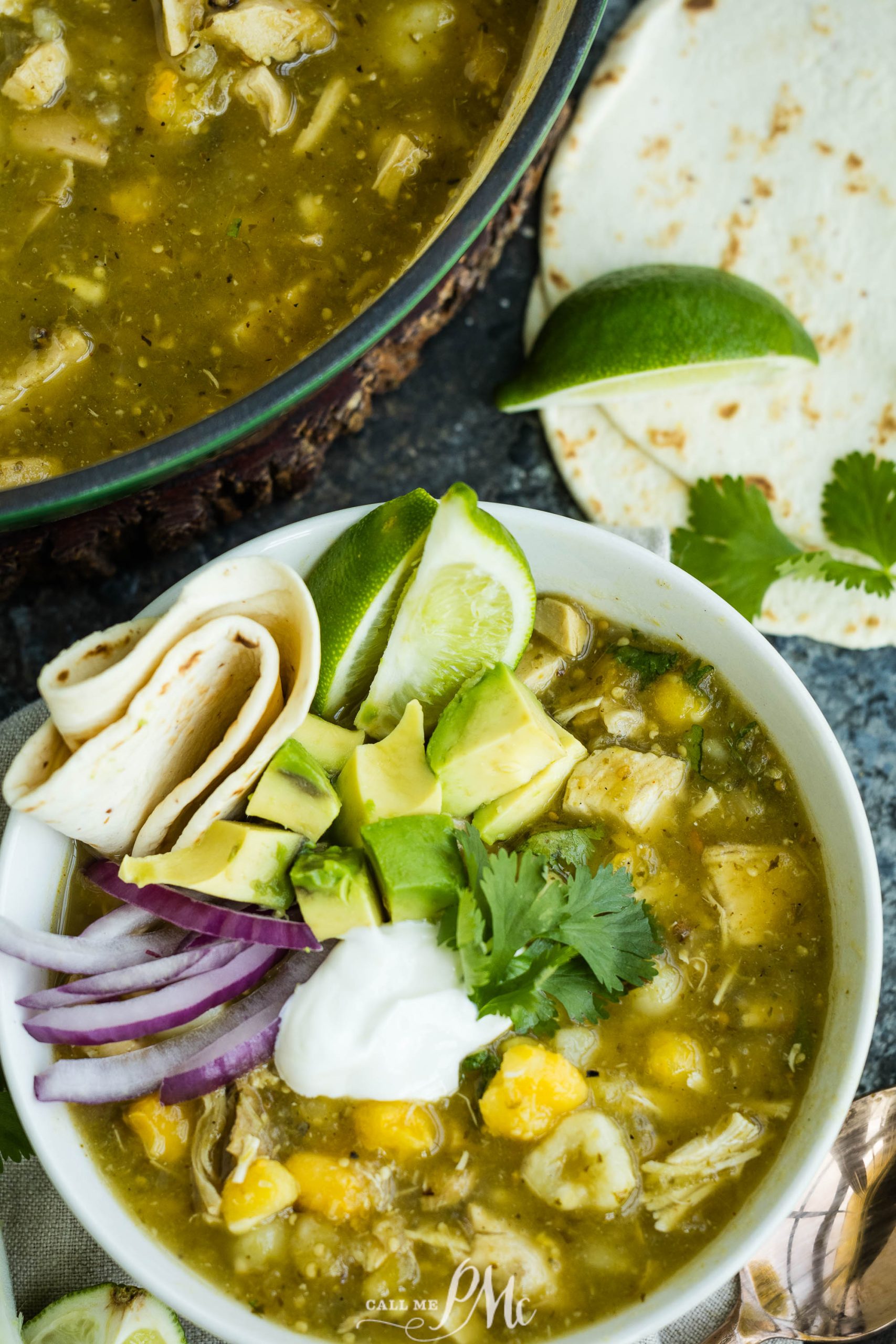 A bowl of green soup with chunks of chicken, avocado, corn, sliced red onions, cilantro, and a dollop of sour cream, garnished with lime wedges and served next to folded tortillas.