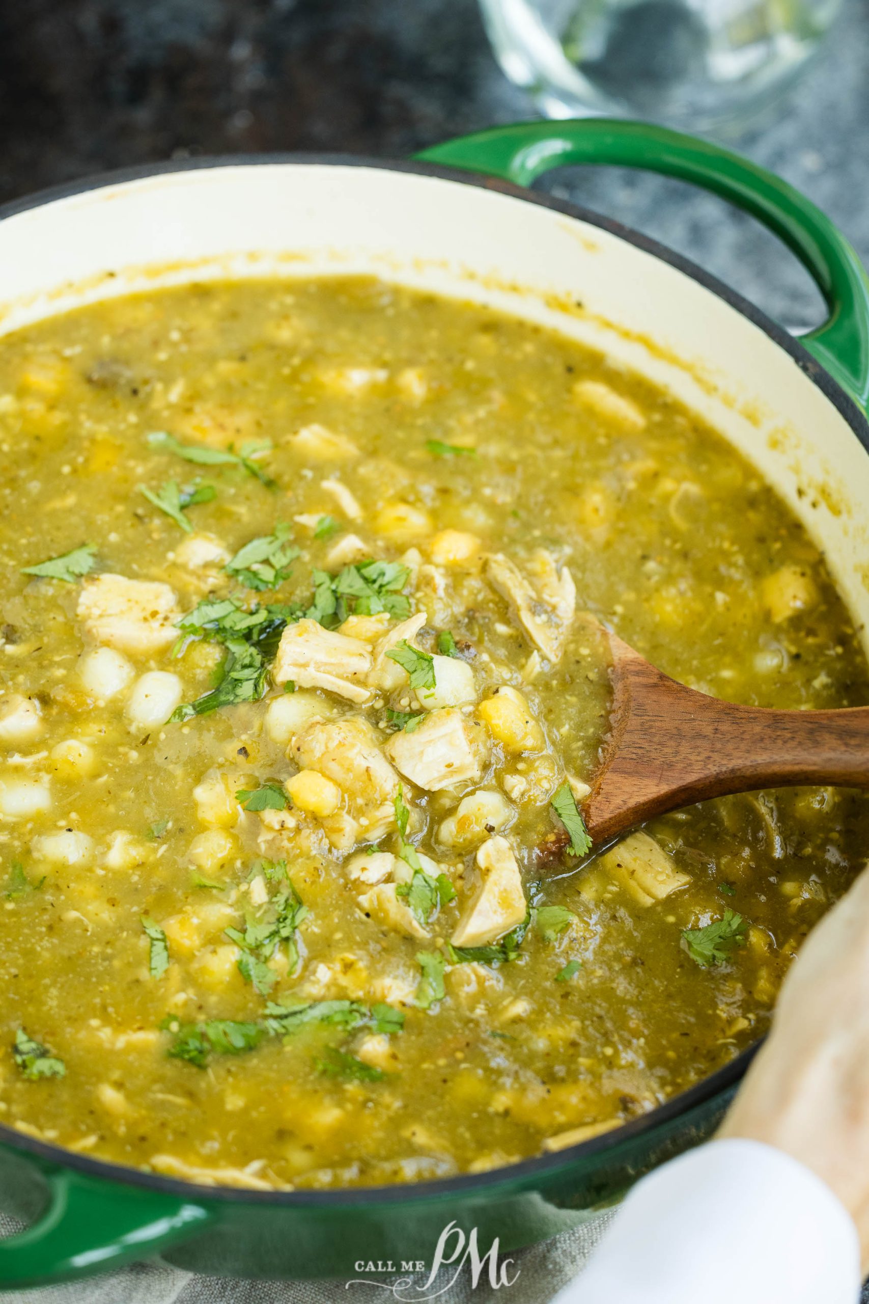 A green pot filled with green chili stew, visible ingredients include corn, chicken, and chopped cilantro, with a wooden spoon stirring the stew.