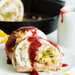 Stuffed turkey breast slices with cranberry sauce on a white plate, with a skillet and sauce pitcher in the background, set on a floral yellow tablecloth.