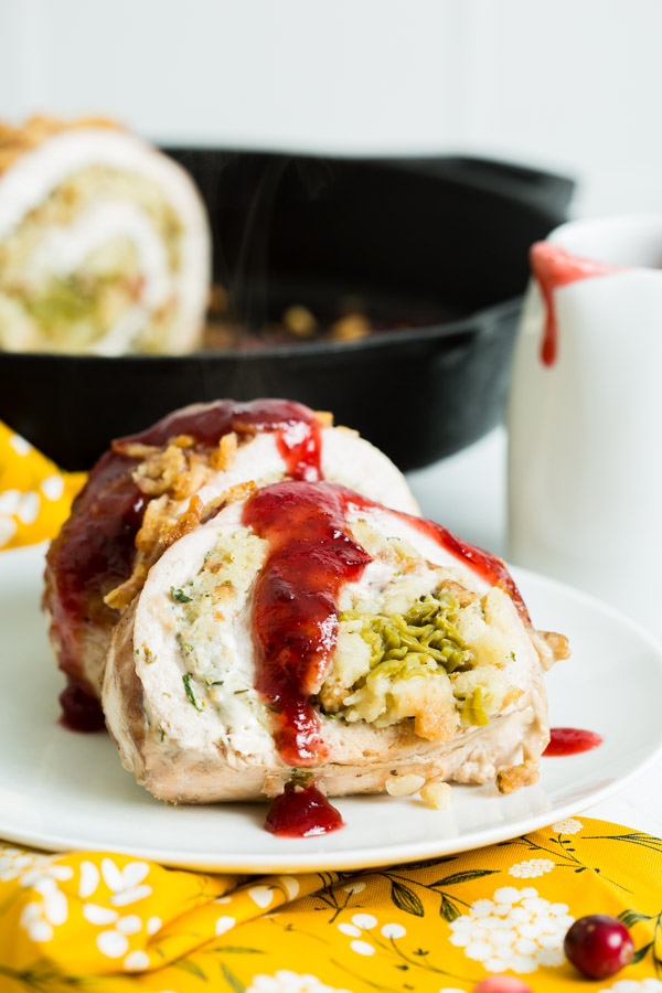 Stuffed turkey breast slices with cranberry sauce on a white plate, with a skillet and sauce pitcher in the background, set on a floral yellow tablecloth.