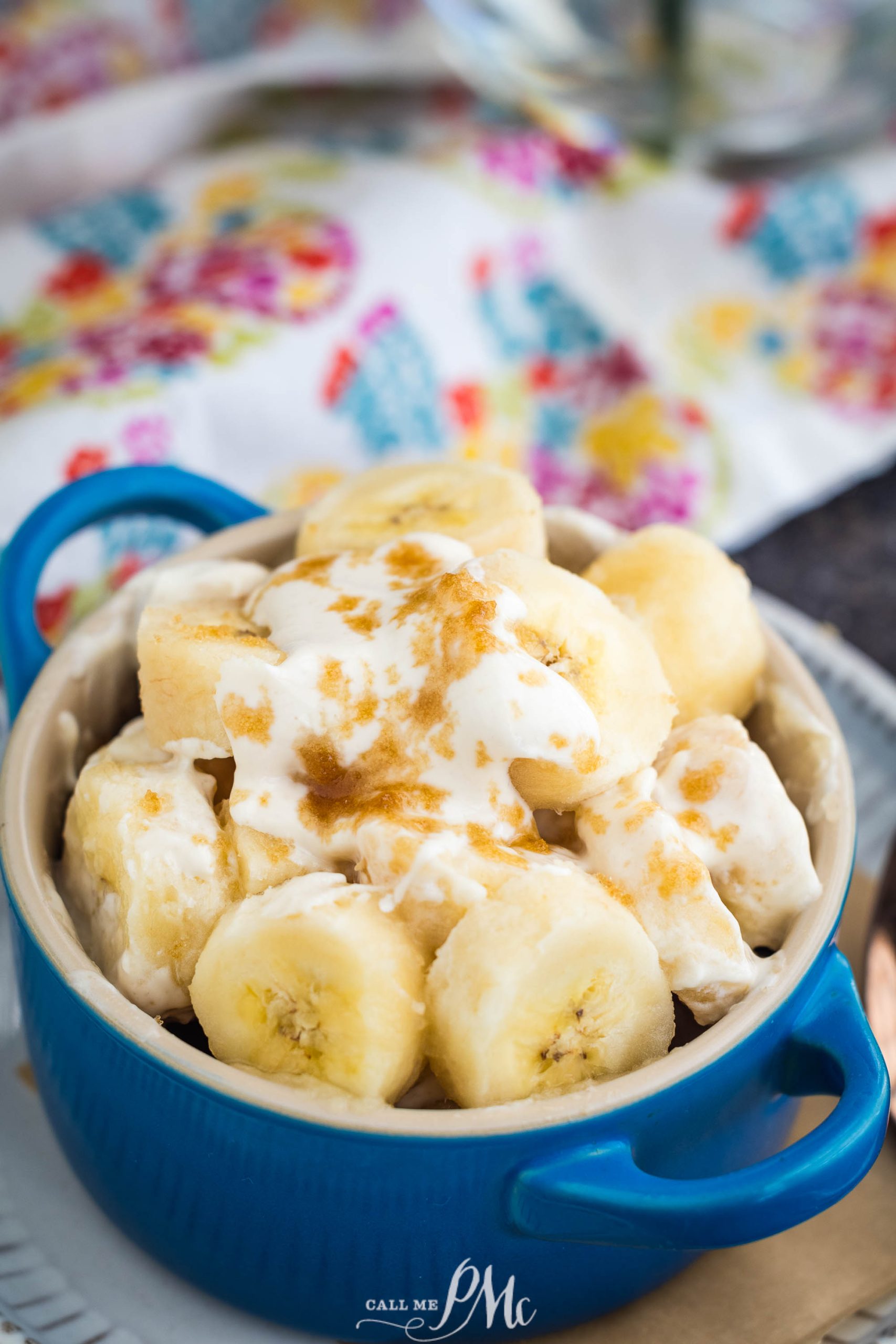 Sliced bananas topped with cream and brown sugar in a blue bowl, on a floral-patterned cloth.