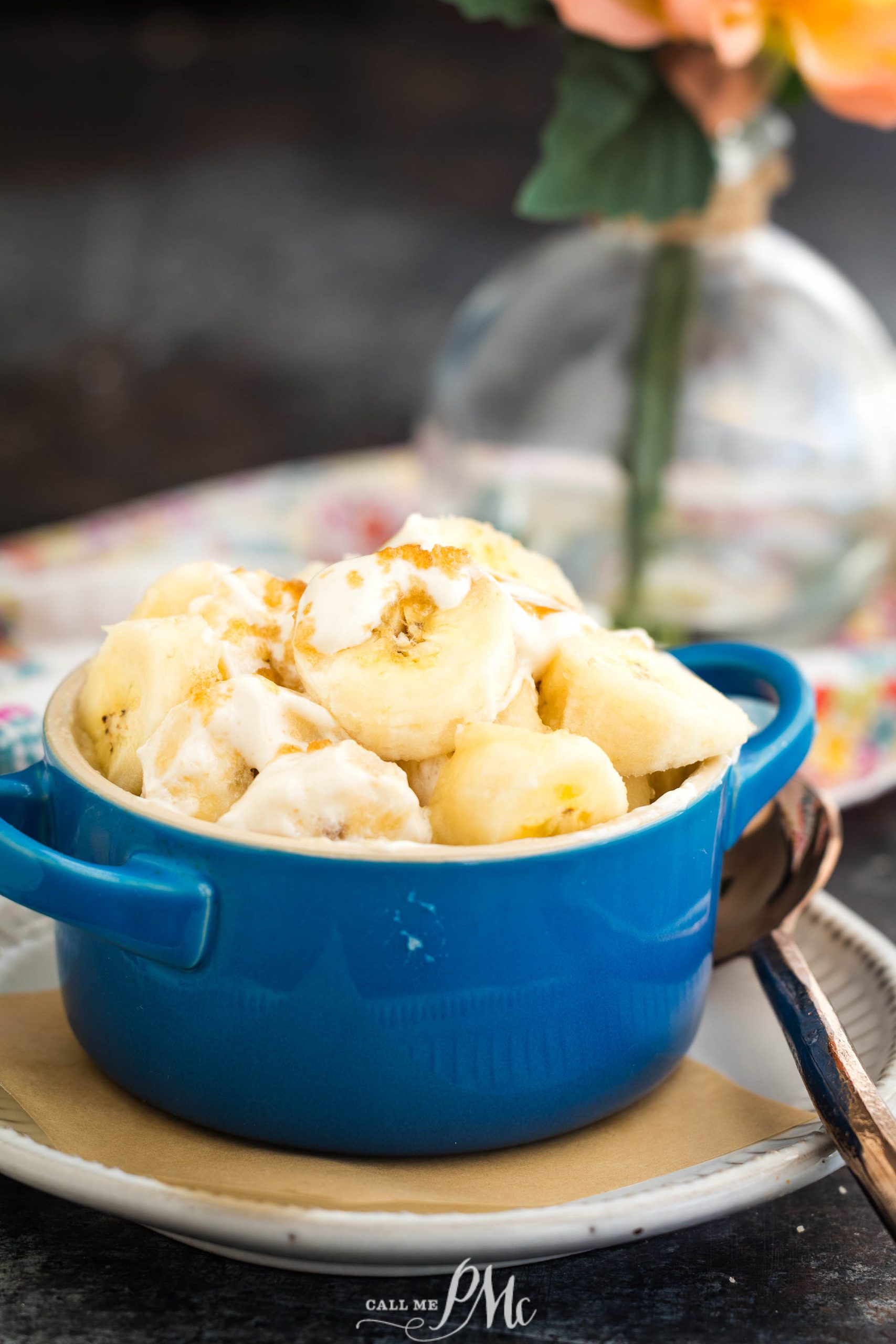 A blue bowl filled with banana pudding topped with crushed cookies, placed on a white plate. A spoon is next to the bowl, with a blurred floral background.