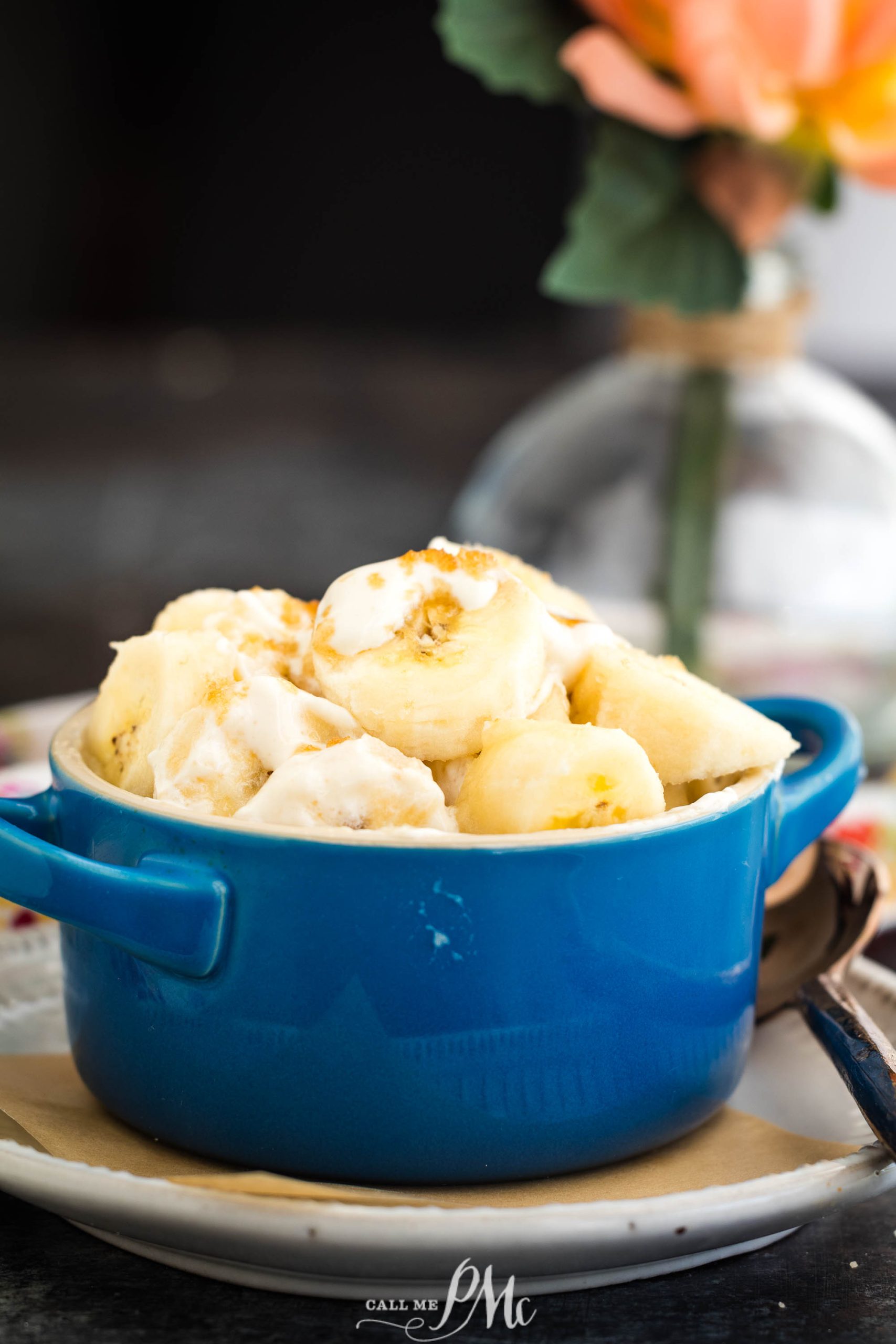 A blue bowl filled with banana slices topped with crushed nuts, placed on a table with a blurred plant in the background.