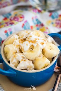 A blue bowl filled with sliced bananas, cream, and a sprinkle of brown sugar, placed on a table with a colorful patterned cloth in the background.