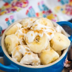 A blue bowl filled with sliced bananas, cream, and a sprinkle of brown sugar, placed on a table with a colorful patterned cloth in the background.