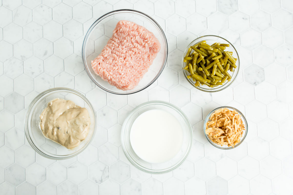 Bowls of ground meat, chopped green beans, creamy soup, milk, and fried onions on a hexagonal tile surface.