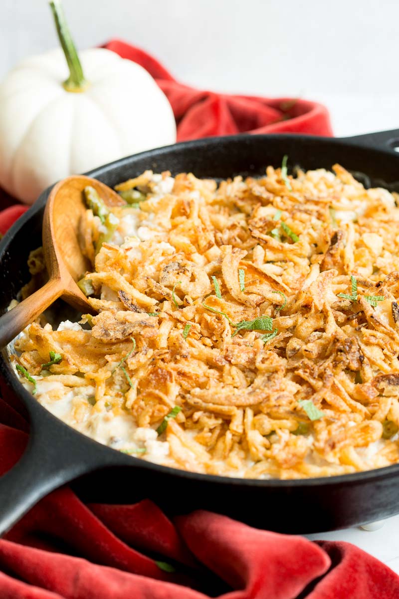 A skillet containing green bean casserole topped with crispy fried onions, next to a small white pumpkin on a red cloth.