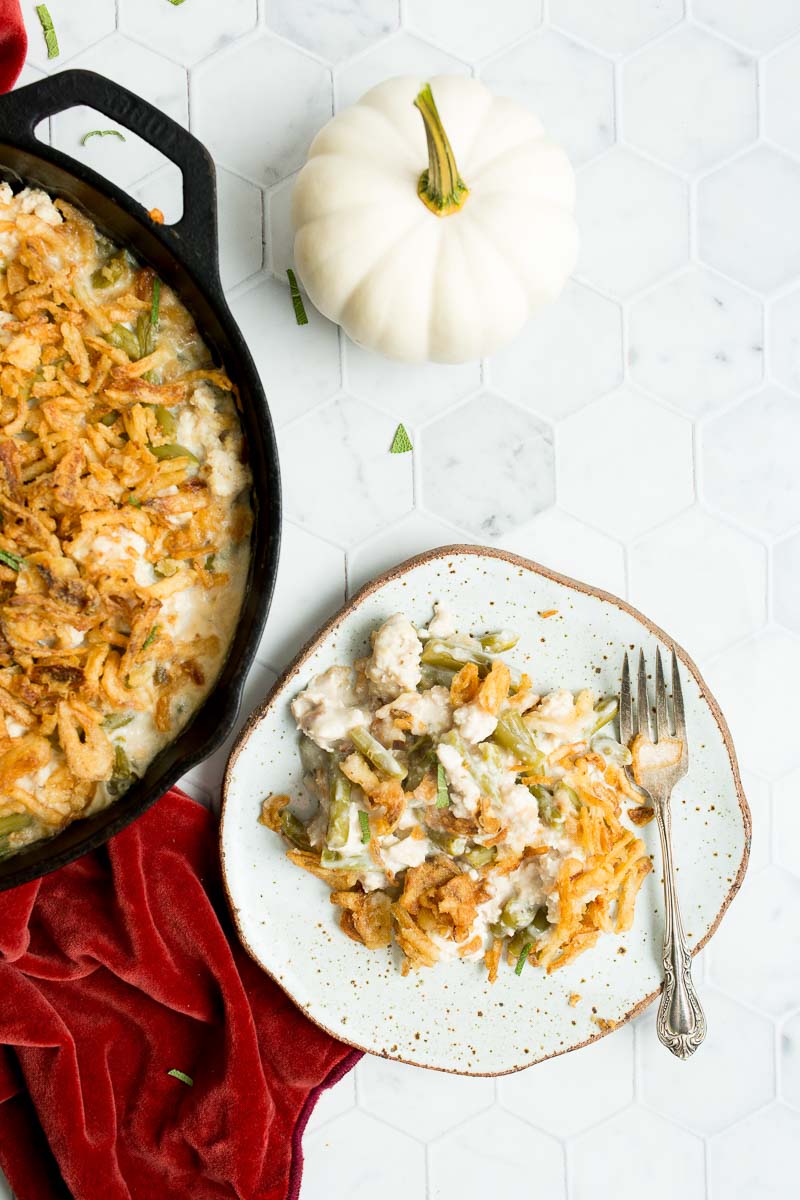 A plate of green bean casserole with a fork next to a skillet, a white pumpkin, and a red cloth on a white hexagonal tiled surface.