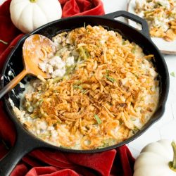 A skillet with green bean casserole topped with crispy onions next to mini white pumpkins and a red cloth.