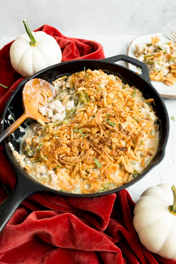 A skillet with green bean casserole topped with crispy onions next to mini white pumpkins and a red cloth.