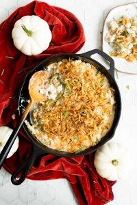 Skillet of casserole topped with fried onions on a red napkin, surrounded by white pumpkins. Plate on the side with a portion served.