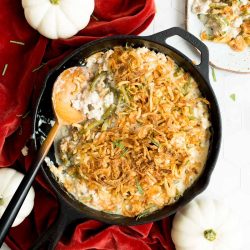 Skillet of casserole topped with fried onions on a red napkin, surrounded by white pumpkins. Plate on the side with a portion served.