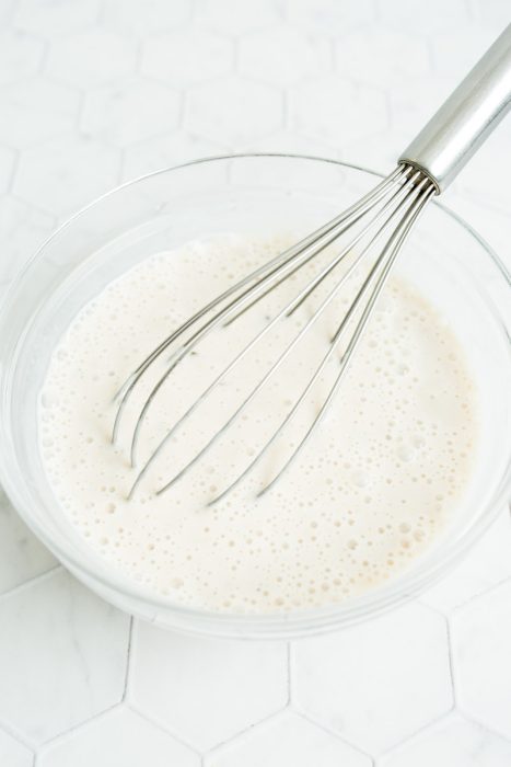 A metal whisk in a glass bowl of frothy batter on a hexagon-patterned surface.